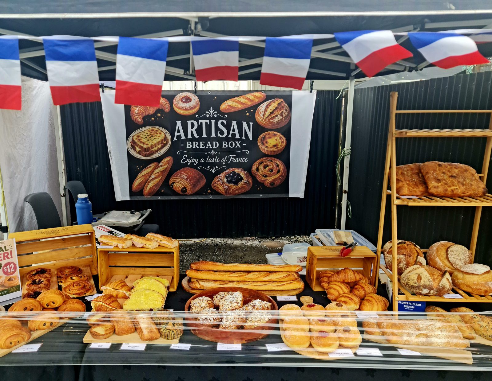 The Artisan Bread Box market stall, decorated with French flag bunting and a banner reading Artisan Bread Box, Enjoy a taste of France, with a full spread of fresh croissants, baguettes, danishes, and sourdough loaves laid out for sale.